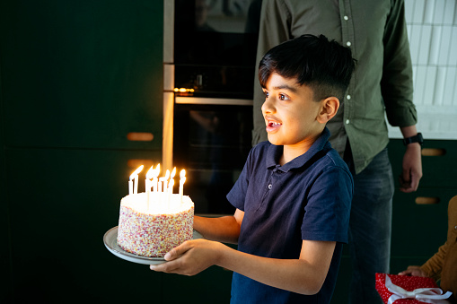Three-quarter front view of 7 year old delivering decorated cake with lit candles to off-camera celebrant as family participates in special day.