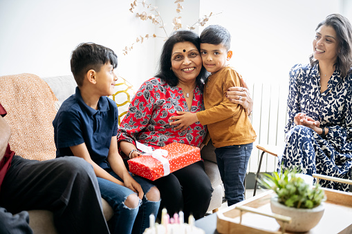 Indian family, including grandsons aged 5 and 7, together in living room, giving gifts, embracing, and smiling at camera.