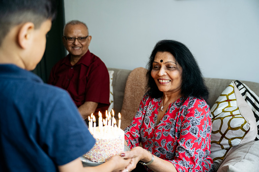 Over the shoulder view with focus on casually dressed woman in late 60s, looking at lit candles and smiling, cheerful senior man in background.