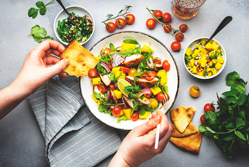 Hands holding fork and tortilla over salad plate with grilled chicken and mango, salsa, tomatoes, cilantro, red onion and lettuce in tex-mex style, gray table background, top view