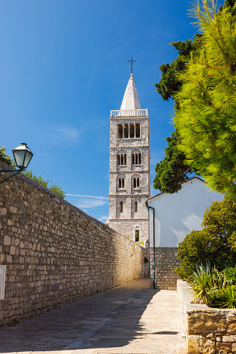 Beautiful view on church tower from a paved alley by the stone wall with lantern, littoral trees in a house garden, Rab town on Rab island in Croatia