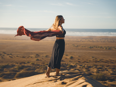 Woman traveler standing on sand with arms outstretched holding a scarf at windy Sahara Desert,Morocco