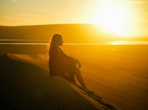 Silhouette of thoughtful woman tourist relaxing on sand dune and admiring beautiful sunset view at Sahara Desert,Morocco