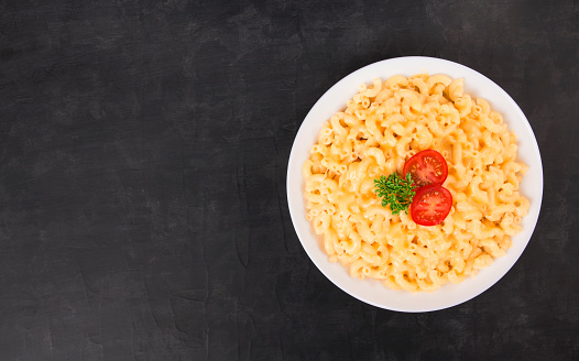 Macaroni and cheese on a white plate with parsley and cherry tomatoes on a black background. American mac and cheese