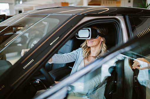 Mid aged woman visits the car dealership, looking at the current offers, talking to the salesperson. Thinking of going EV or staying with the internal combustion car. She is trying out the VR presentation of the car.