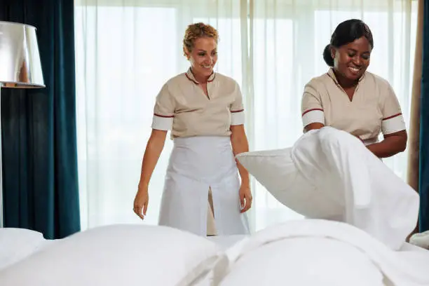 Two diverse hotel maids in white uniforms laughing while cleaning the guestroom together