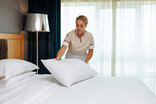 Young uniformed Caucasian maid putting the clean pillows on the bed