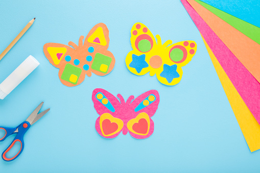 Butterfly shapes from colorful paper on light blue table background. Pastel color. Making decoration elements. Closeup. Top down view.