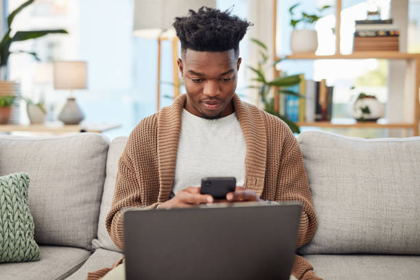 istock Laptop, phone and man on a sofa networking on social media. mobile app or the internet. Technology, computer and young African male person browsing online with a cellphone in his living room at home. 1502313659