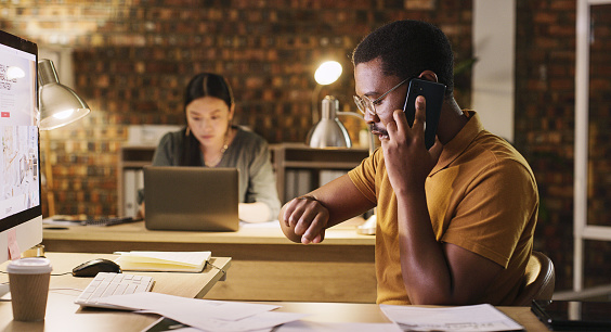 Time, late and a black man on a phone call at work for business, networking and deadline. Talking, browsing and an African employee working overtime on a computer while speaking on a mobile at night