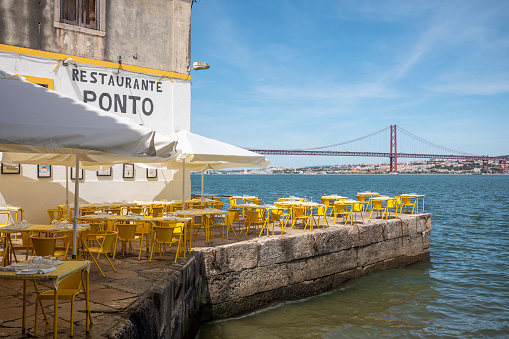 Lisbon, Portugal - May 25, 2022: Restaurant with yellow chairs and tables overlooking Ponte 25 de Abril in Lisbon Portugal at day.