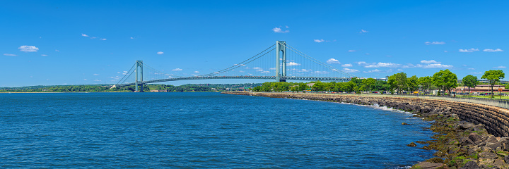 High resolution stitched Panorama of the Verrazano-Narrows Bridge on a spring morning with New York Bay Bank Reinforced with Rocks and Cars driving on the Belt Parkway. The bridge connects boroughs of Brooklyn and Staten Island in New York City. It was built in 1964 and is the largest suspension bridge in the USA. Historic Fort Wadsworth is next to the bridge foot. Canon EOS 6D full frame censor camera. Canon EF 85mm F/1.8 lens. 3:1 Image Aspect Ratio. This image was downsized to 50MP. Original image resolution is 79MP or 15356 x 5119 px