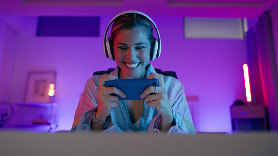 Happy woman, mobile gaming or game console with headphones, neon lights or streamer at night. Female gamer playing esports, online or tech connection for digital streaming on application at dark home