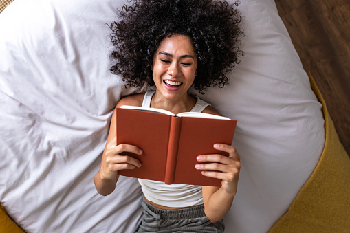 Top view of young happy multiracial woman lying on the bed reading a book laughing. Lifestyle concept.