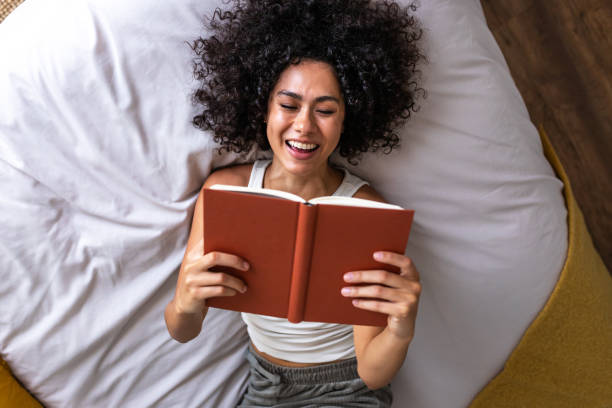 Top view of young happy multiracial woman lying on the bed reading a book laughing.