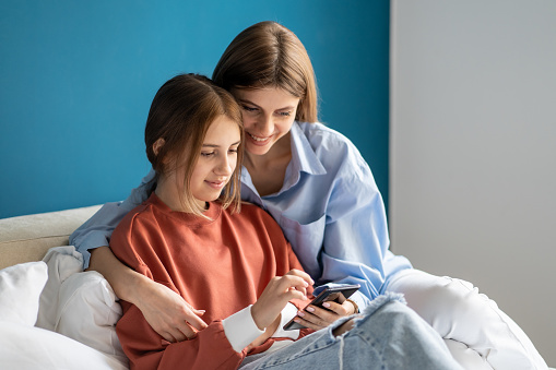 Positive mom daughter watching funny video in smartphone together sitting in bed at home. Family woman teen girl browsing mobile phone searching information. Leisure, digital recreation, shopping.