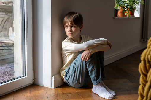 Kid psychological trauma. Lonely child boy sadly look to window hug knees sits on floor at home alone. Upset offended child thinking about family problems, bad relationship, failures with school peers