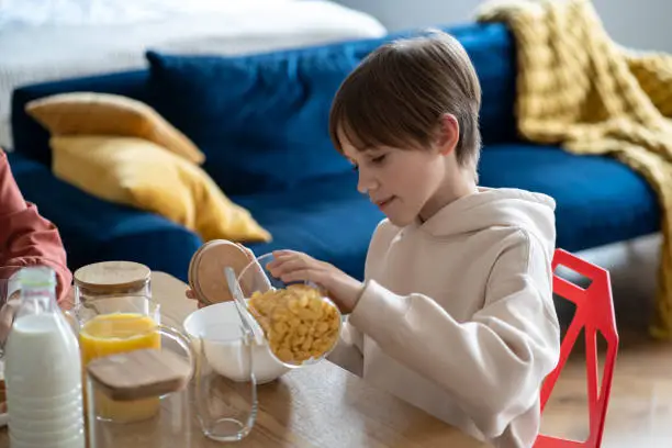 Photo of Teen boy preparing breakfast for himself pouring cornflakes in bowl sitting at table on kitchen.