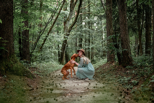 Young woman ang Hungarian Vizsla dog standing on the footpath in the forest in a summertime.