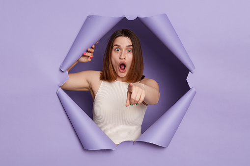 Shocked surprised brunette woman breaking through purple paper hole wearing casual white top pointing at camera choosing you keeps mouth open.