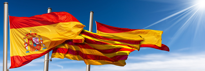 Close-up of Spanish and Catalan flags (la Rojigualda and Senyera) with flagpole, blowing in the wind on a blue sky with clouds, sunbeams and copy space.