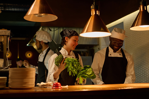 Young smiling woman in uniform bringing fresh broccoli and green leafy vegetables to chef chopping ingredients for salad or some other dish