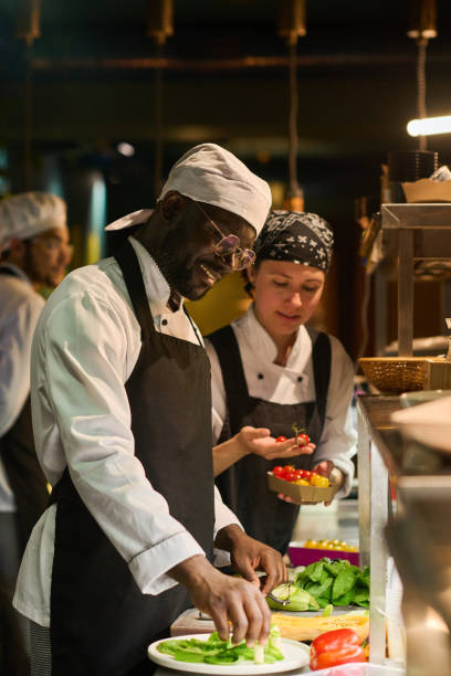 Happy young African American male chef mixing fresh ingrdients on board Happy young African American male chef in uniform mixing fresh ingredients on small round board before putting it into pan commercial kitchen photos stock pictures, royalty-free photos & images