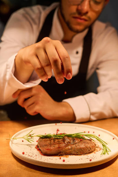 istock Focus on hand of young man in uniform sprinkling beef steak with spices 1502384698