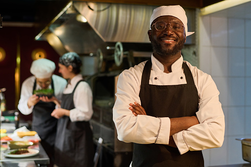 Happy African American male chef in uniform standing in front of camera in restaurant kitchen and keeping his arms crossed by chest