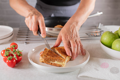 Fresh pan fried ham and cheese sandwich is cut by woman hand in half on a plate on kitchen counter background. Closeup, front view