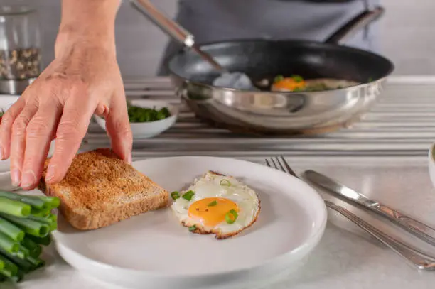 Woman with apron is preparing breakfast. She is serving a slice of toast on a plate with fresh fried egg, sunny side up. Closeup, front view