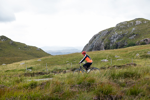 A senior multicultural female submersed in nature. She is riding her bike down a hill in Torridon, in the Scottish Highlands on an overcast day. There is a dramatic green backdrop around her.