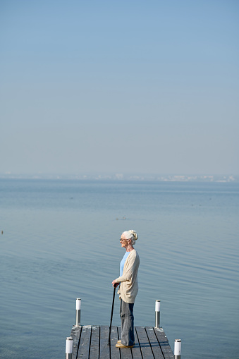 Long shot of retired woman with walking cane standing on pier by large blue lake and enjoying beautiful view while having promenade