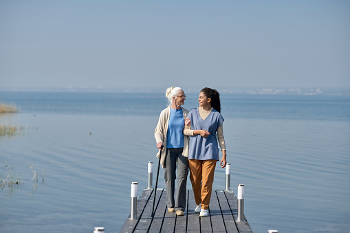 Long shot of aged woman and her granddaughter walking along pier against blue lake and sky and looking at one another while chatting