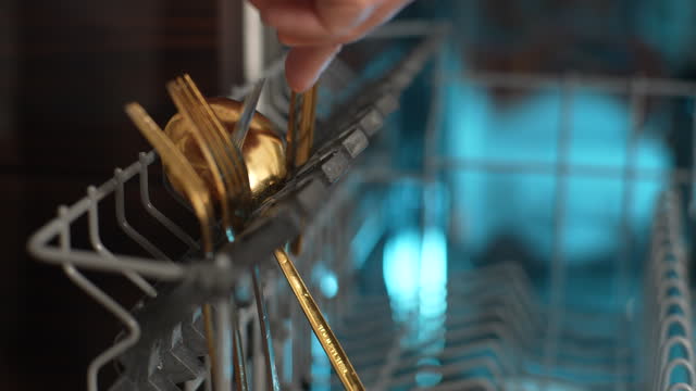 Close-up of unrecognizable man preparing basket and loading backlit dishwasher machine with dirty stainless flatware. Concept using modern kitchen appliance