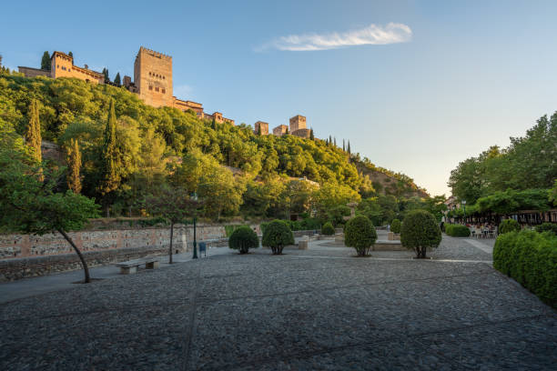 In the photo, the brilliance of Granada's Spanish architecture is evident under the sunny sky. The intricate patterns adorning the buildings' exteriors stand out crisply against the azure backdrop. The sunlight bathes the stone structures, bringing out their inherent beauty and craftsmanship. Notably, the distant silhouette of the Alhambra adds an air of mystery and historical significance, enhancing the scenic charm nestled amidst the mountainous landscape.