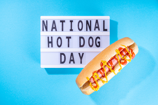 National Hot Dogs day background, hotdog summer party festival foods, Two tasty classic american hot dogs with sauces and lightbox sign with inscription National Hot Dog Day on blue background