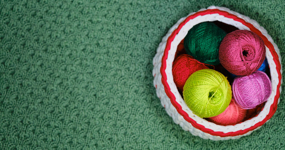 Yarn for crocheting. Top view, web banner with copy space. Concept hobby of handmade needlework from home. Several woolen multicolored balls of thread in wicker basket on a green crocheted background.