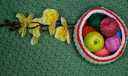 Yarn for crocheting. Top view, web banner with copy space. Several woolen multicolored balls of thread in wicker basket on green crocheted background. Yellow branch of orchid flower nearby.