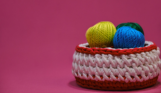 Concept craft handmade needlework hobby from home. Yarn for crocheting. Several woolen multi colored balls of thread in a wicker basket on a pink background. Side view, web banner with copy space.