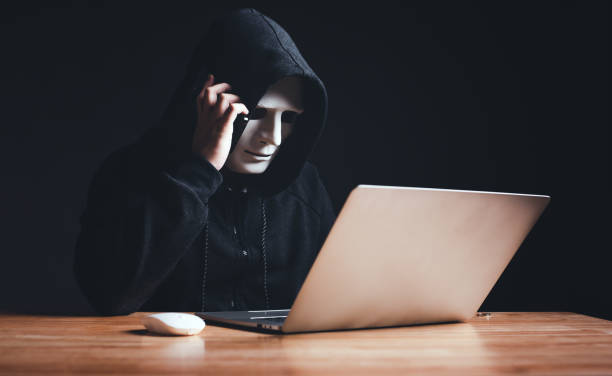 Black hat hacker in hood using laptop computer and call smartphone to victim on desk hacking privacy sensitive data hack in dark room background. Cyber security cyber crime concept. Hacking phishing stock photo