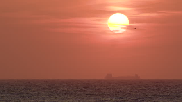Closeup view of cargo bulker on its transportation voyage during sunset. A huge white disk of the rising sun against the background of the sky and clouds painted in red and orange colors