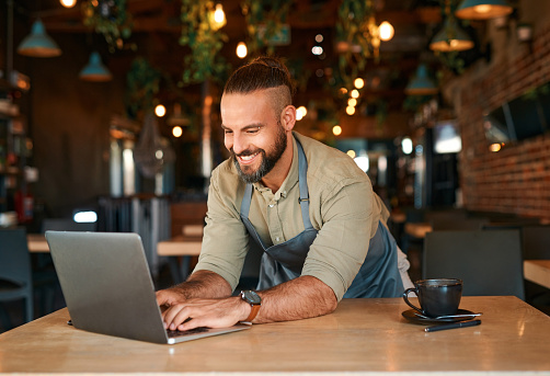 Small business, owner and happy man at laptop in cafe for inventory management, restaurant planning or food industry. Male entrepreneur working on computer in retail coffee shop, startup and internet