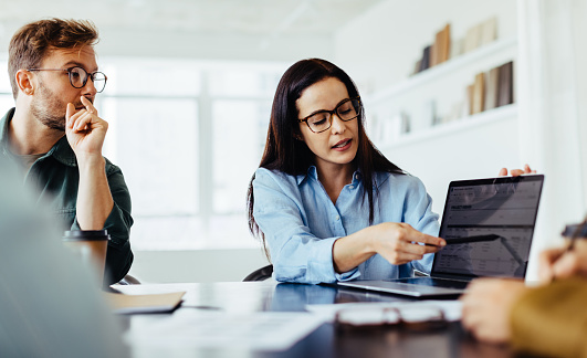 Business woman discussing a project report with her team in an office. Female professional using a laptop while leading a meeting with her colleagues.