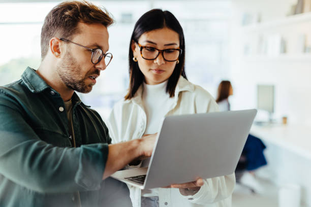 Design professionals using a laptop together in an office Design professionals standing in an office and using a laptop together. Two young business people discussing a project. coworker stock pictures, royalty-free photos & images