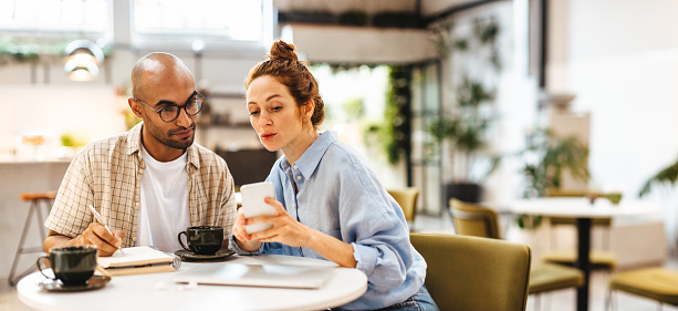 Business woman sharing her mobile phone with her client while discussing work in a meeting. Caucasian female entrepreneur collaborating with her male associate in a cafe.