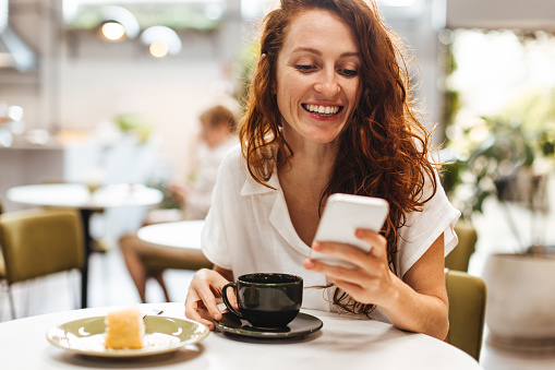 Young woman browsing her favourite social media apps on her smartphone as he enjoys a solo coffee date in a cafe. Caucasian woman with ginger hair enjoying her own company in a coffee shop.