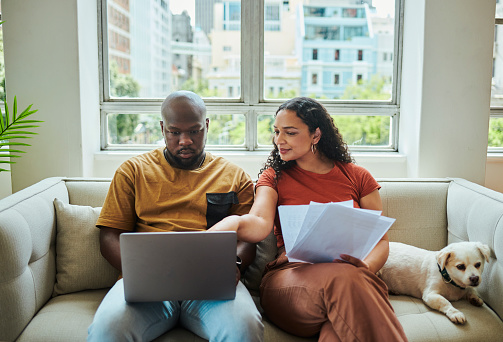 A young interracial couple working on their budget and finances on a sofa from home with their dog using a laptop computer, stock photo
