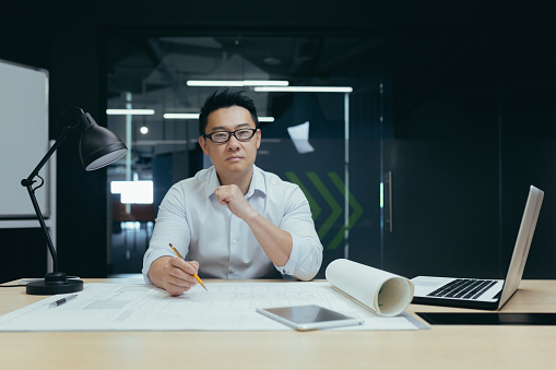 Portrait of a young Asian male designer, architect, engineer looking confidently and seriously at the camera, working in the office at a table with papers and projects.