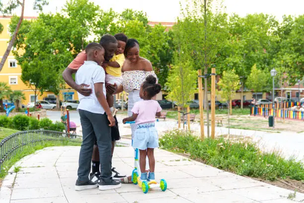 A beaming African family embraces in a city park: father cradles a toddler, mother stands beside an 11-year-old son, and a little girl glides on a scooter towards them.
